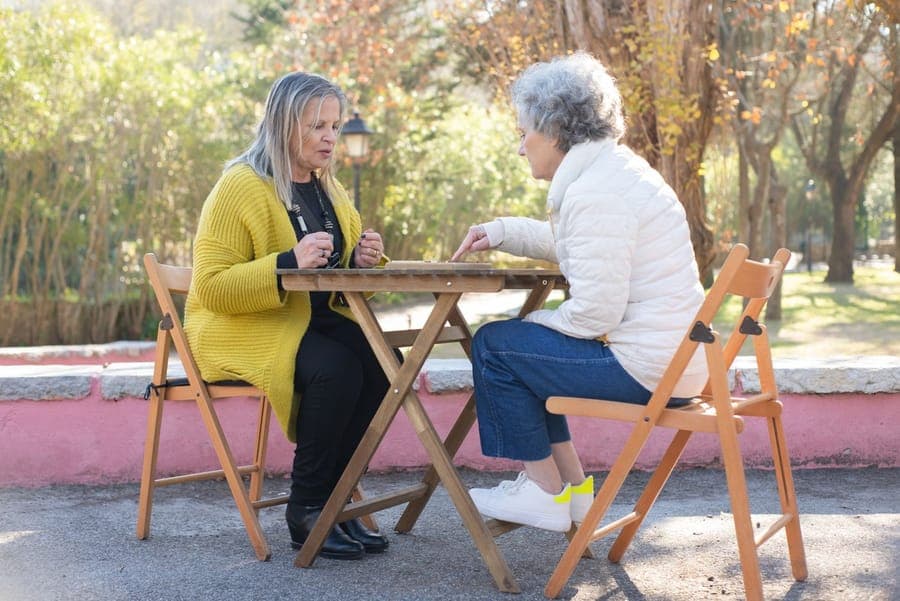 Two older women chatting over a board game at a table in a sunny park